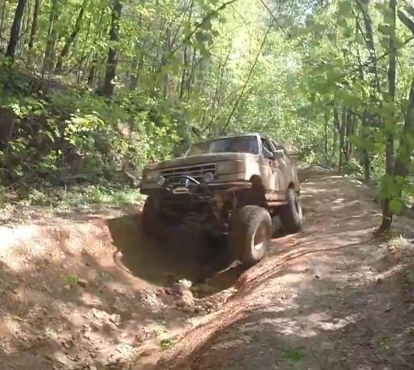Lifted 1988 Ford Bronco driving through a muddy trail in a forest.