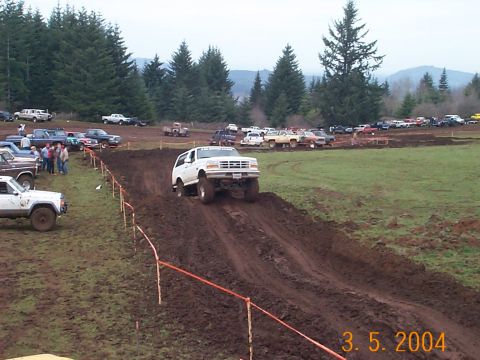 1983 Ford Bronco - A white SUV navigates a muddy track during an off-road event with other vehicles in the background.