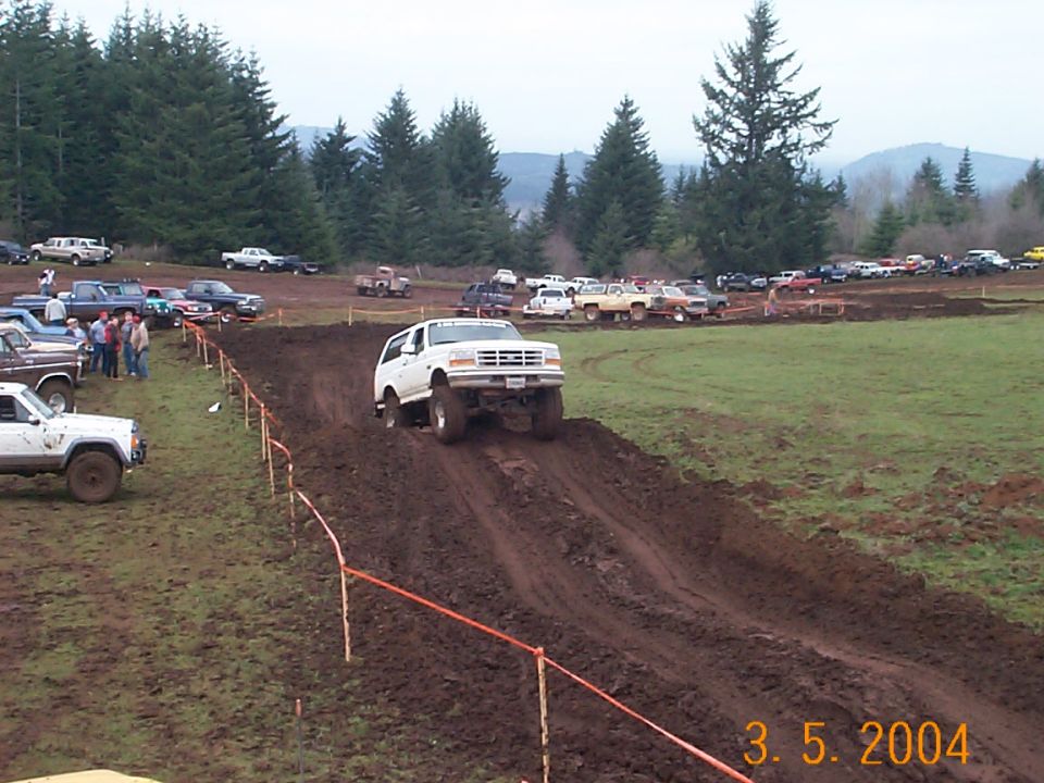 White lifted Ford 4x4 climbing rutted mud hill at off-road event, spectators and trucks in background