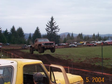 1983 Ford Bronco - A brown pickup truck soars through the air during a mud bog event, with spectators and other vehicles in the background.