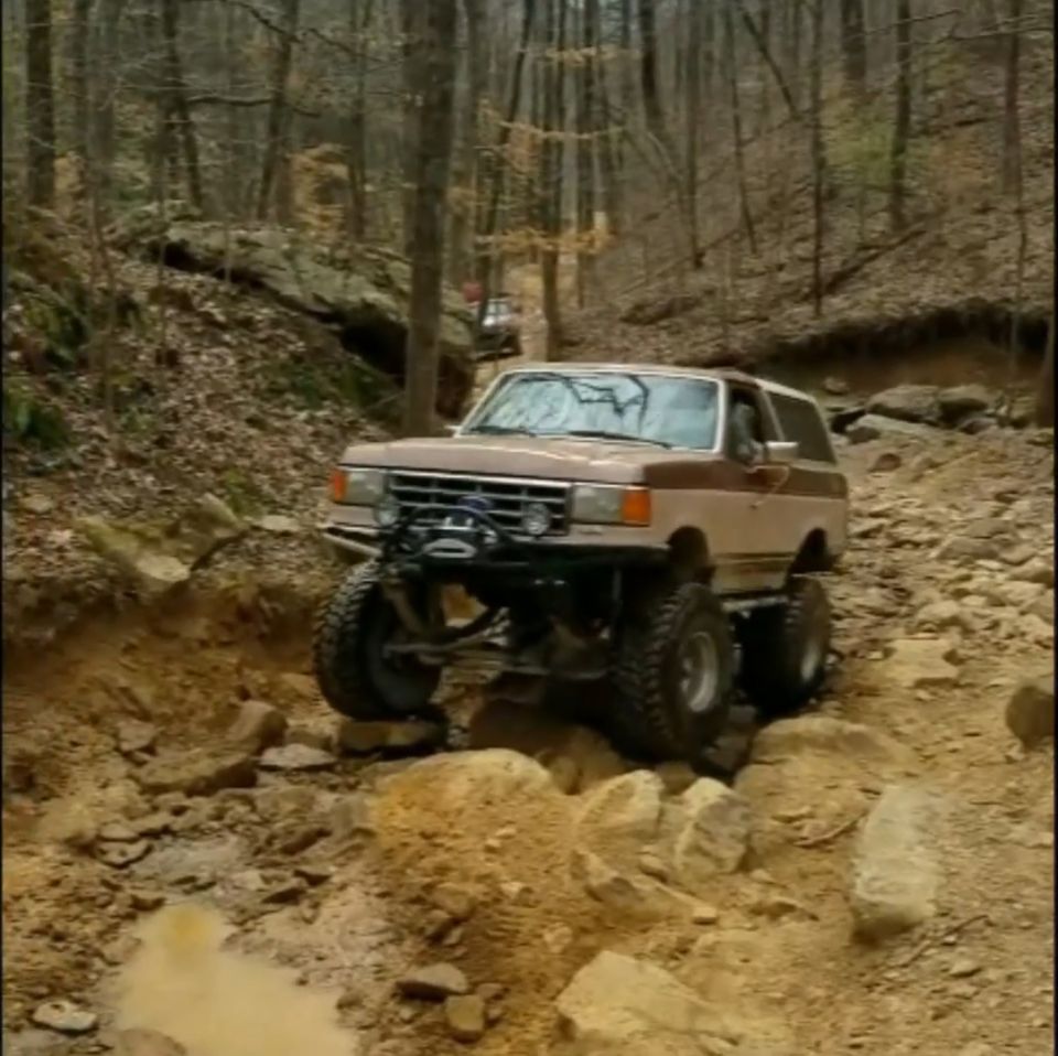 Lifted 1988 Ford Bronco with large tires and winch on a rocky trail in the woods.