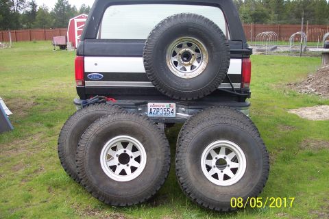 1989 Ford Bronco - The rear of a black Ford Bronco with a spare tire mounted on the tailgate and four additional tires with silver rims laid out on the grass…