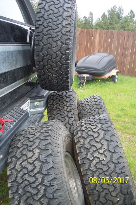 1989 Ford Bronco - A spare tire is mounted on the back of a dark-colored SUV, with four additional tires stacked on the ground in front of it. A small utility…