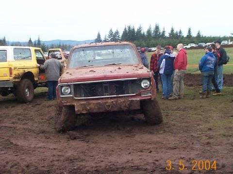 1983 Ford Bronco - Muddy red pickup truck stuck in the mud with a yellow SUV nearby and spectators watching.