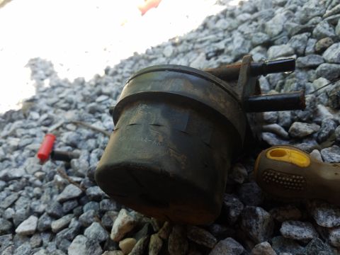 1988 Ford Bronco - A close-up, low-angle shot shows a black automotive part, possibly a fuel filter or vacuum canister, resting on a bed of grey gravel. The p…