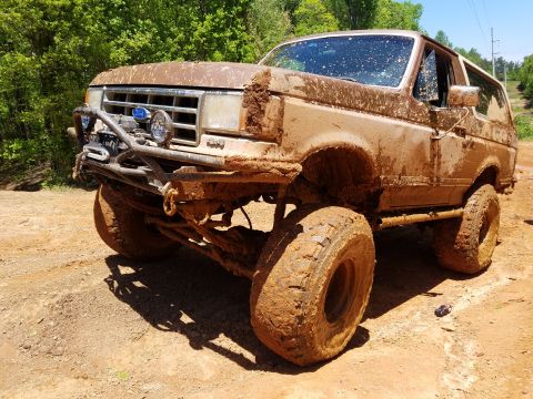 1988 Ford Bronco - A heavily mud-covered Ford Bronco is parked on a dirt path surrounded by trees. The off-road vehicle features large tires, a custom front b…