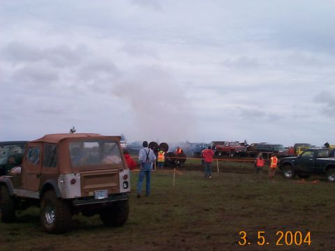 1983 Ford Bronco - Several off-road vehicles are gathered in a muddy field under a cloudy sky, with some people observing.
