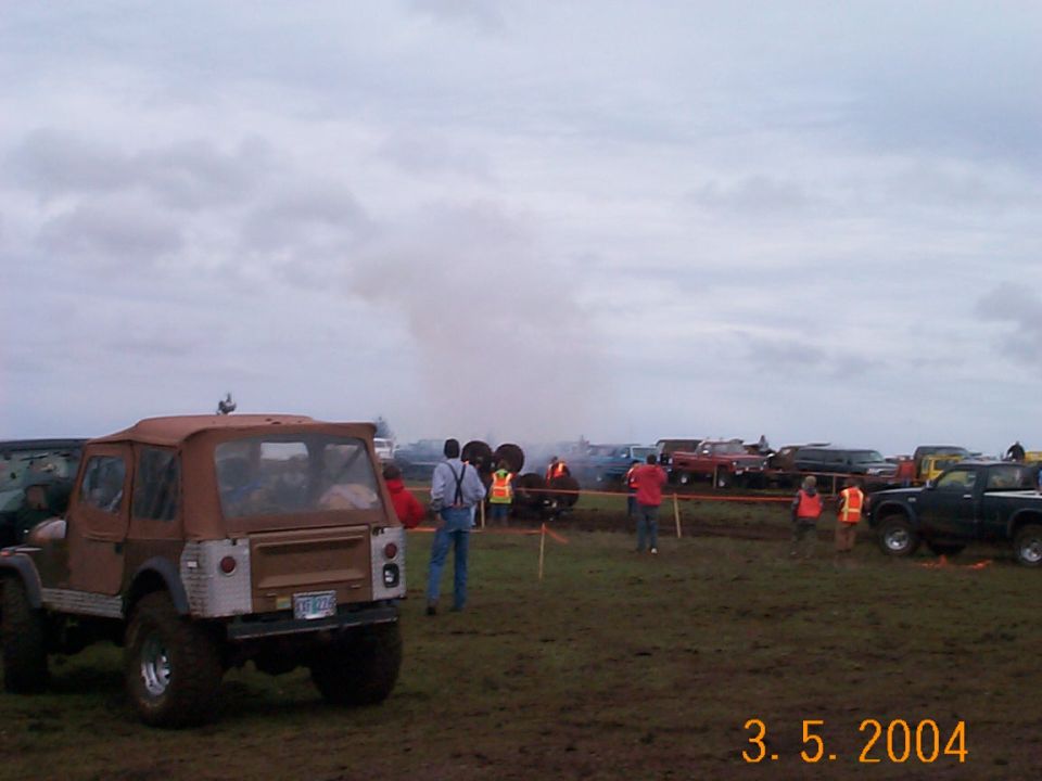 Tan soft-top Jeep parked at muddy off-road event with vehicles and spectators in background