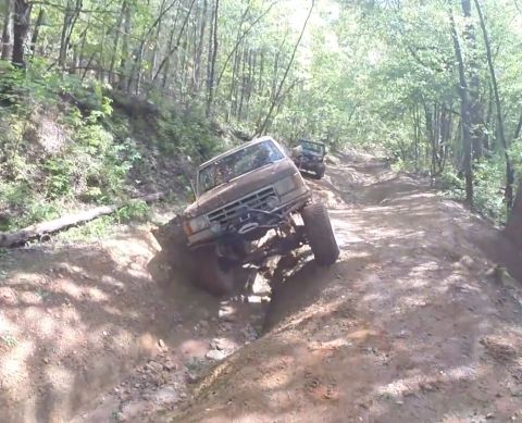 1988 Ford Bronco - A brown 1988 Ford Bronco is navigating a steep, muddy off-road trail in a forest. The vehicle is tilted to the side as its front tire climb…