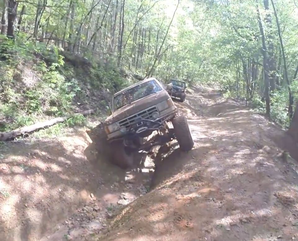 1988 Ford Bronco off-roading on a steep, muddy trail in a forest.