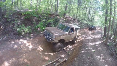 1988 Ford Bronco - A tan, lifted Ford Bronco is navigating a muddy, wooded trail. Another dark-colored off-road vehicle is visible further up the path.