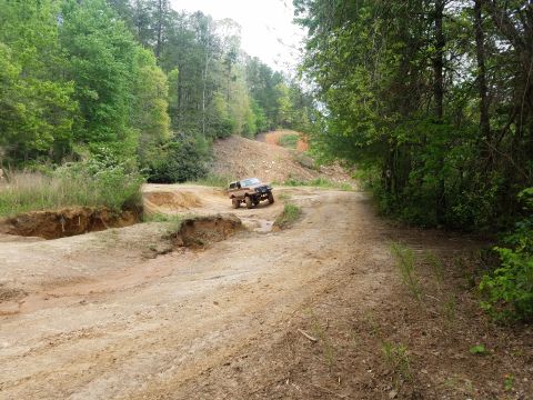 1988 Ford Bronco - A tan 1988 Ford Bronco is navigating a muddy, off-road trail with steep dirt banks on the left side of the frame. The vehicle is angled tow…