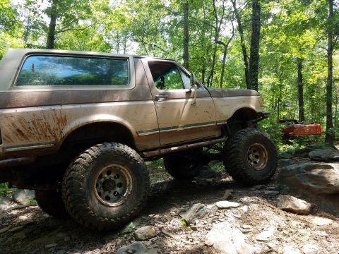 1988 Ford Bronco - This lifted SUV navigates a challenging rocky trail surrounded by trees.
