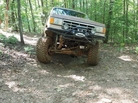 1988 Ford Bronco - Ford Bronco navigating a wooded trail with aggressive off-road tires and a custom bumper.