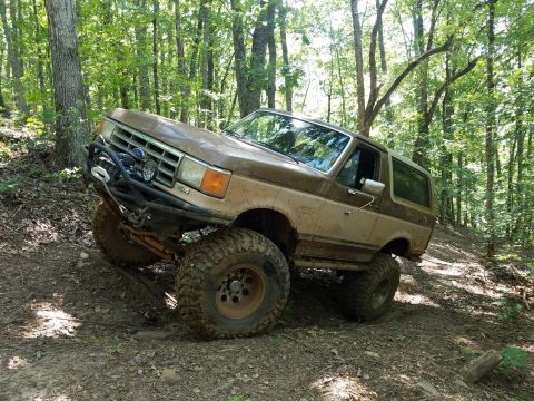 1988 Ford Bronco - A lifted Ford Bronco navigates a wooded trail with large tires.