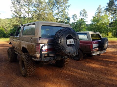 1988 Ford Bronco - Two Ford Broncos are parked on a dirt road with trees in the background. The front vehicle has a spare tire on its tailgate.