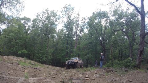 1988 Ford Bronco - A tan Ford Bronco navigates a challenging off-road course surrounded by dense woods.
