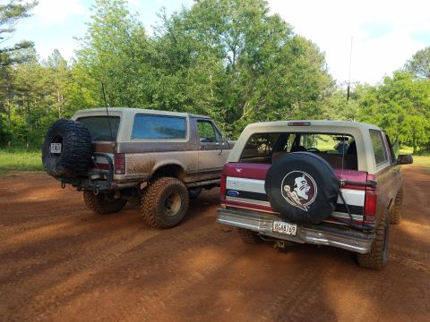 1988 Ford Bronco - Two Ford Broncos are parked on a dirt road, one brown and one red, with a spare tire cover on the red one.