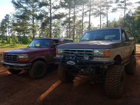 1988 Ford Bronco - Two Ford Broncos, one brown and one red, are parked side-by-side in an outdoor setting.