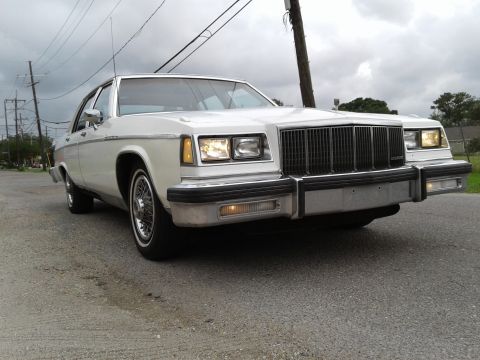 1984 Buick Electra - A white sedan with a prominent vertical grille and wire wheels is parked on a gravel road under a cloudy sky.