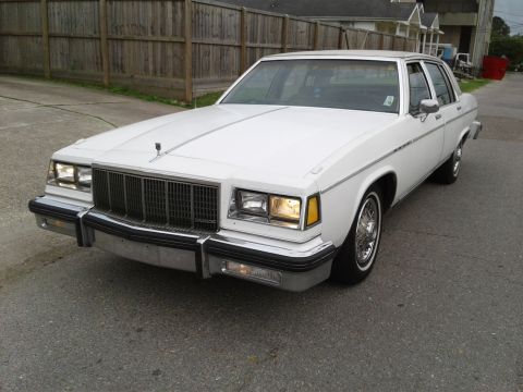 1984 Buick Electra - A white sedan is parked on a street with a wooden fence in the background.