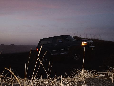 1996 Ford Bronco - A Ford Bronco sits on a hill as the sun sets, with tall grass in the foreground.