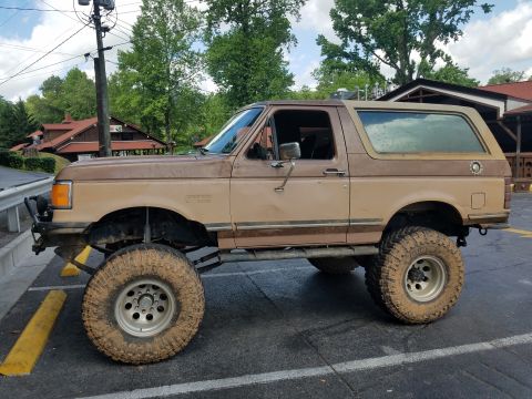 1988 Ford Bronco - This lifted Ford Bronco is parked in a lot with buildings and trees in the background.