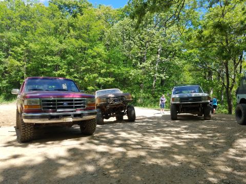 1988 Ford Bronco - Several Ford trucks are parked on a dirt road surrounded by trees.