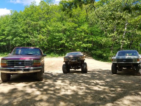 1988 Ford Bronco - Three lifted trucks, including two Ford Broncos and a Ford F-series, are parked on a dirt road surrounded by trees.