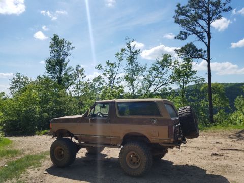1988 Ford Bronco - A lifted brown Ford Bronco sits on a dirt road with trees and a blue sky in the background.
