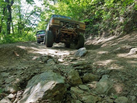 1988 Ford Bronco - Two off-road vehicles navigate a challenging rocky trail through a wooded area.
