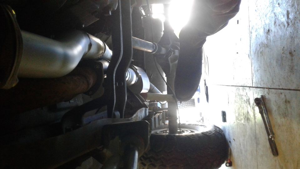 Underside view of a vehicle's exhaust system and suspension components. A person is working on it
