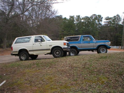 1978 Ford Bronco - Two vintage Ford trucks parked side by side on a gravel/grass area, both facing right in profile. In front is a white full-size Bronco SUV…