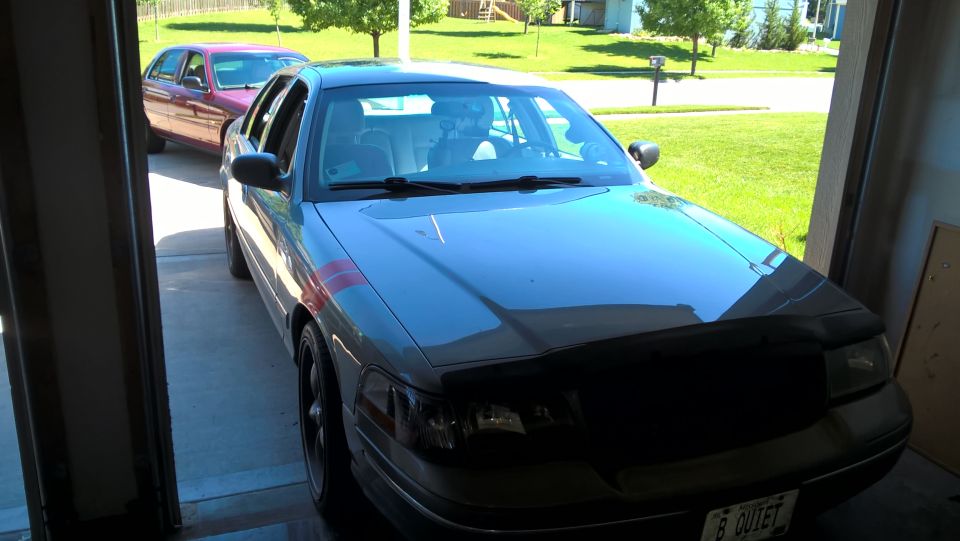 Silver Ford Crown Victoria with red stripes and black hood bra in a garage.
