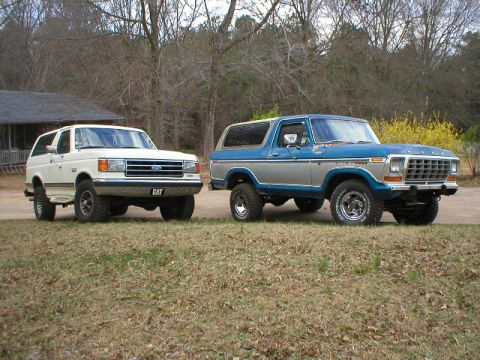 1978 Ford Bronco - Two full-size Ford Bronco SUVs parked on a paved driveway; on the right, a lifted two-tone blue and silver Bronco on large tires, and on th…