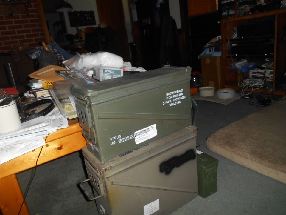Two green ammo cans stacked, with a table and mattress in a cluttered room.
