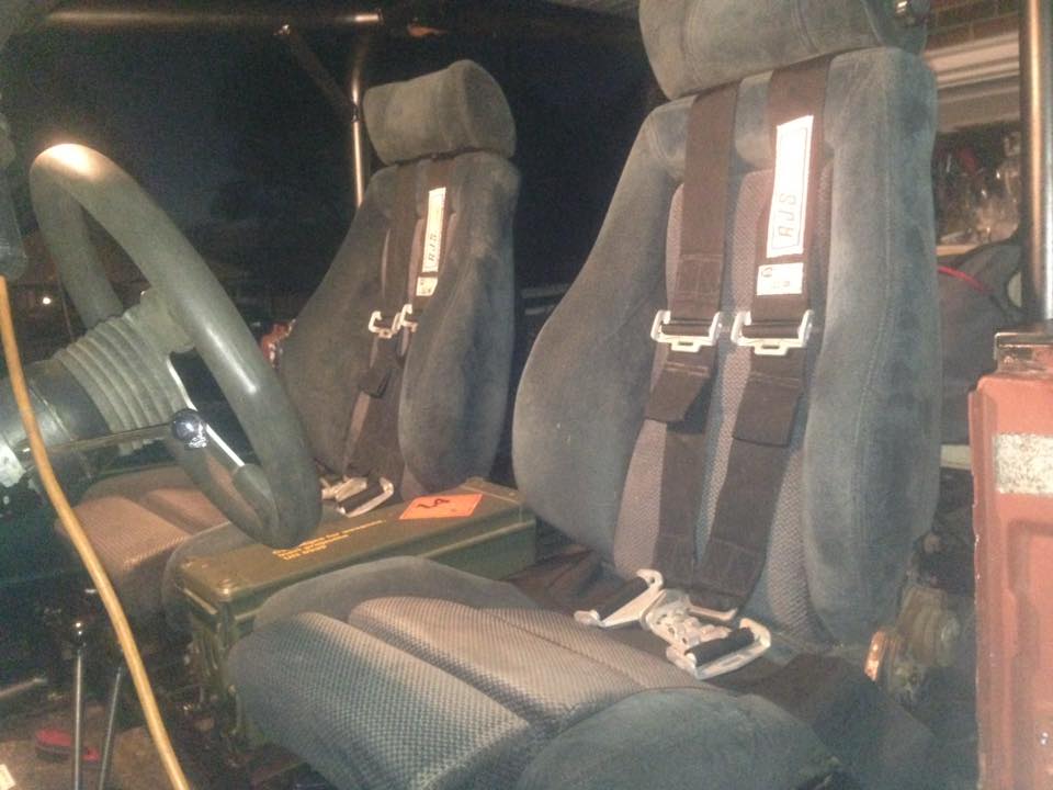 Interior of a vehicle with bucket seats, racing harnesses, steering wheel, and ammo can.