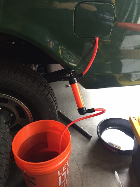 1978 Ford Bronco - A manual fluid transfer pump is shown siphoning liquid from a dark green vehicle's fuel tank into an orange bucket. A black oil drain pan i…