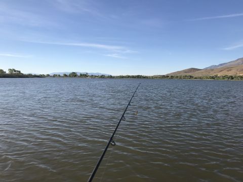1997 Dodge 3500 - A fishing rod extends into a calm lake with a distant shoreline and mountains under a clear blue sky.