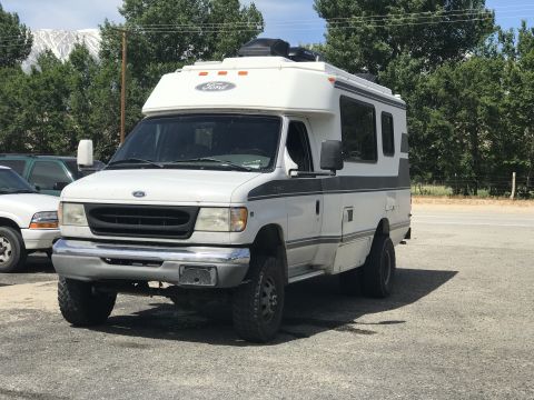 1997 Dodge 3500 - A white Ford E-Series RV with off-road tires and a dark grey stripe is parked on a gravel lot with a mountain and trees in the background.
