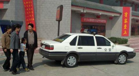1998 Ford Crown Victoria - A white and grey sedan with a light bar on the roof is parked outside a building with red banners.