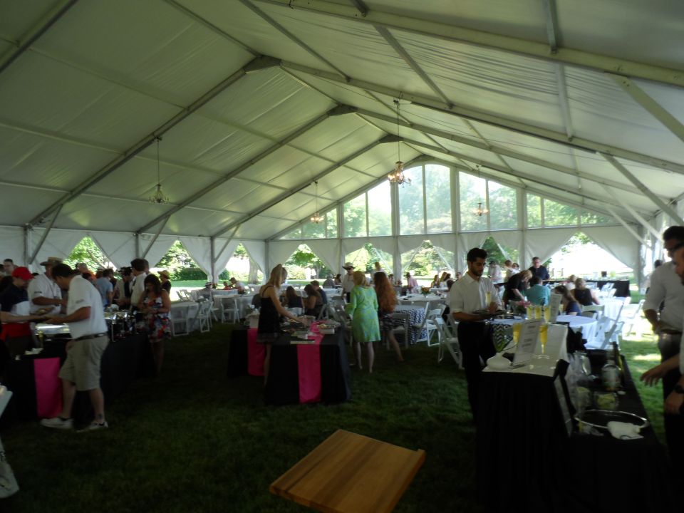 Outdoor event under a tent with people gathered around food and drink tables.