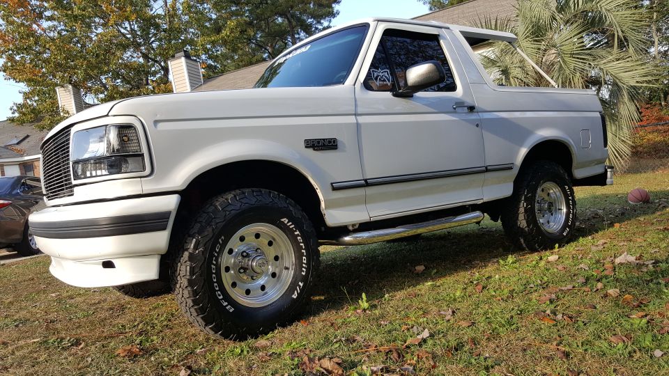 White 1990s Ford Bronco with aftermarket wheels and chrome side step parked on grass.