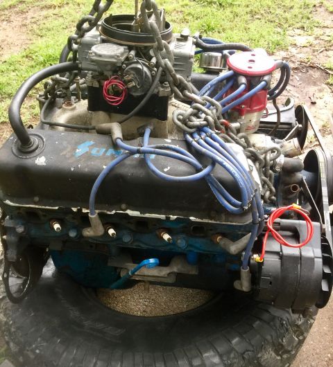 1974 Ford Bronco - Engine assembly, likely a V8, is hoisted by chains and rests on a large tire. Blue spark plug wires and a red distributor cap are visible.