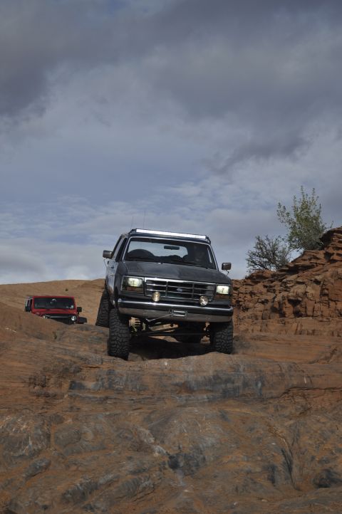 1991 Ford Bronco - Two off-road vehicles navigate a , rocky landscape under a cloudy sky.