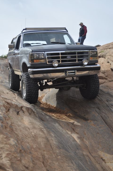 1991 Ford Bronco - A modified Ford Bronco navigates a challenging rocky trail, with a person observing from atop the vehicle.