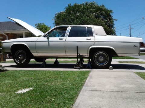 1984 Buick Electra - A white sedan is raised on jack stands for maintenance, with its hood open under a clear blue sky.