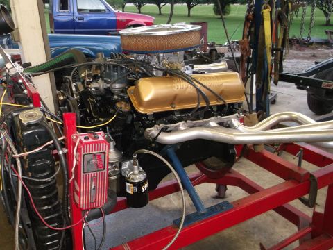 1964 Ford Galaxie - A V8 engine with gold valve covers and custom headers is mounted on a red stand, with parts of two pickup trucks visible in the background.