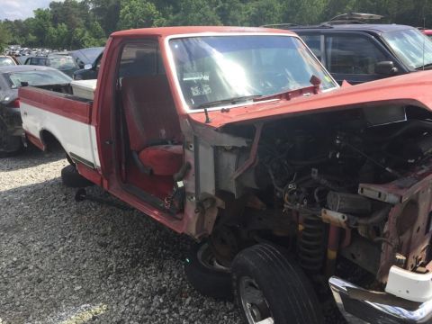 1988 Ford Bronco - This red and white pickup truck, likely from the late 1980s, is seen in a junkyard with its front end heavily damaged and removed.