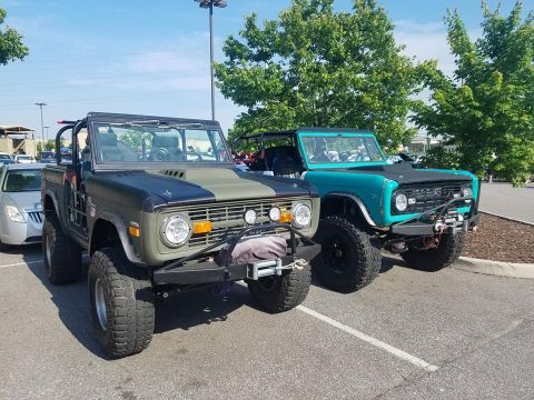 1972 Ford Bronco - Two Ford Broncos are parked in a lot, one with a two-tone paint job and the other teal.